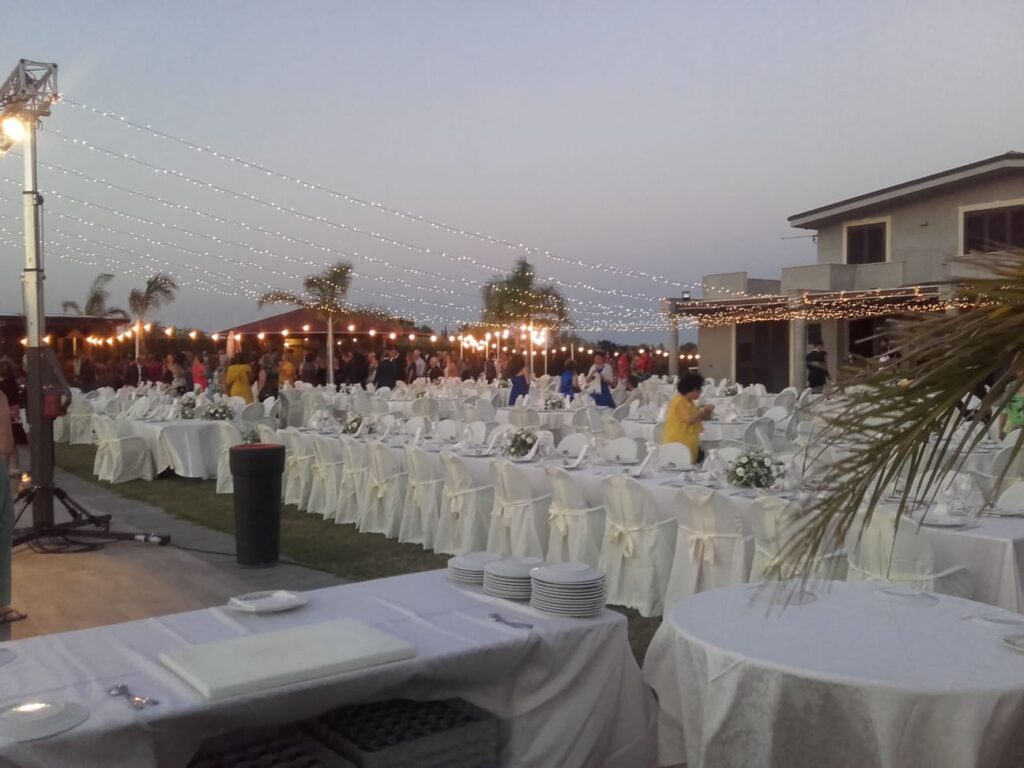 Allestimento catering in giardino con tavoli rotondi e sedie rivestite, sotto alberi di ulivo, con camerieri in servizio
