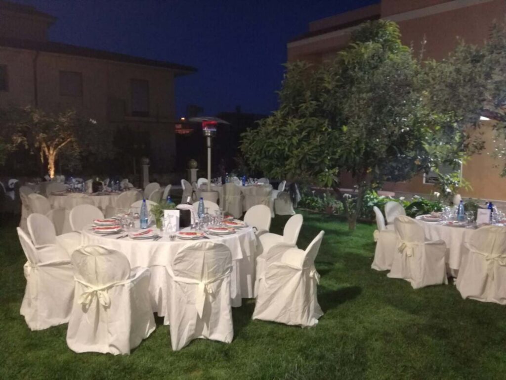 Allestimento catering in giardino con tavoli rotondi e sedie rivestite, sotto alberi di ulivo, con camerieri in servizio